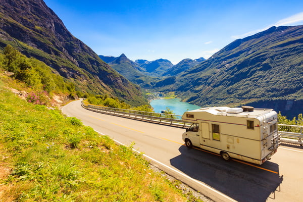 An RV is traveling down the road with a view of a crystal blue lake in the background. The lake is surrounded by mountains and valleys.