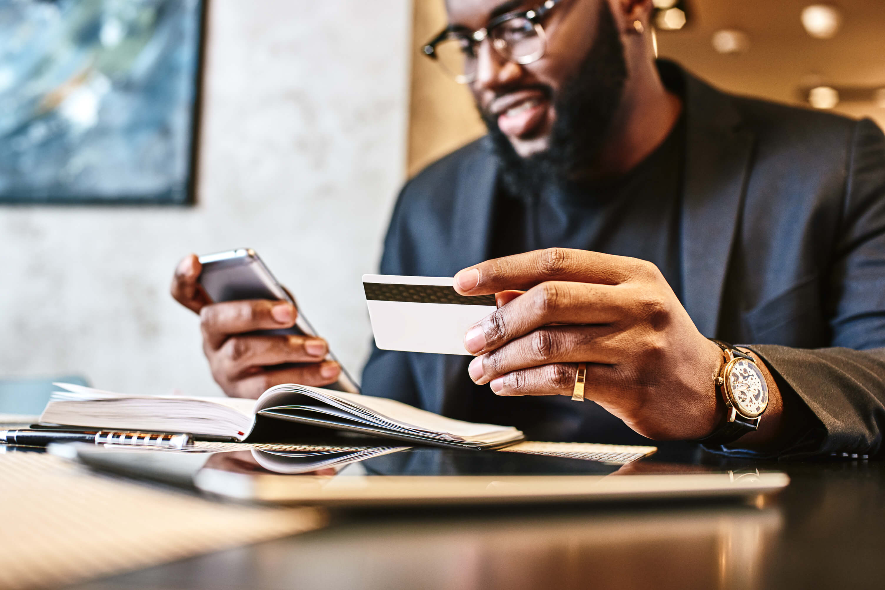 A well dressed man sits at a table with an open book in front of him, a cell phone in his right hand, and a credit or debit card in his left hand.