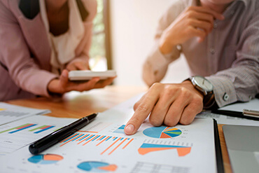 A close up image of some charts and graphs with a man’s hand pointing at the papers and a woman holding a calculator and leaning over the desk.