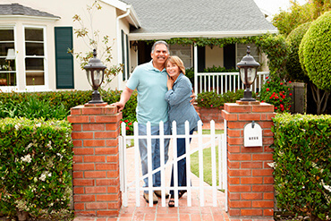 An older couple standing behind a white picket gate, between two brick pillars, in front of their home.