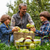 A person and two boys sitting in the grass with a basket of apples