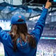 A sports fan watching her favorite team in a blue jacket and hat from Fanatics with her arms raised in the air