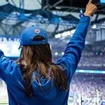A sports fan watching her favorite team in a blue jacket and hat from Fanatics with her arms raised in the air